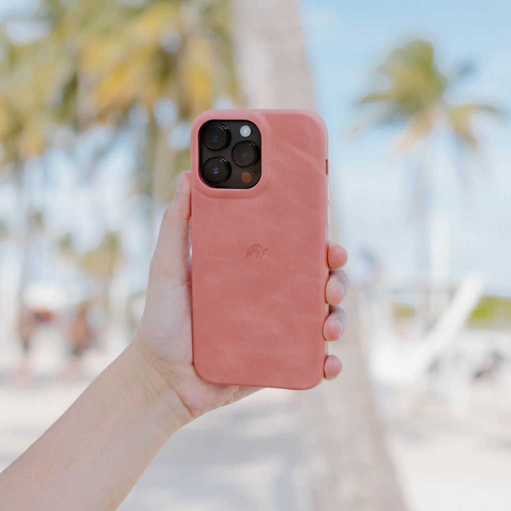 A person holding a pink Minimalist Case for iPhone 16 in a beach setting with palm trees in the background.