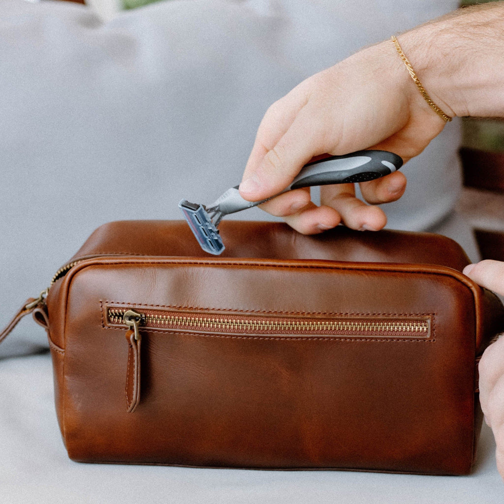 A person holding a razor above a brown leather Dopp Kit bag.